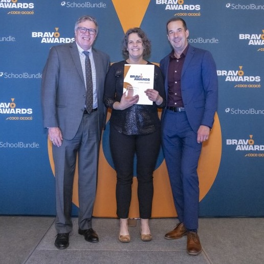 Jesse Green stands on stage between two presenters, holding a certificate at the 2024 BRAVO Awards ceremony. A step-and-repeat banner with the BRAVO Awards and SchoolBundle logos is visible behind them..